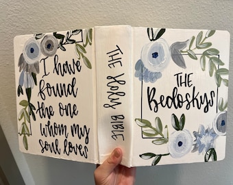 May include: A white book with a floral design and the text "THE Holy BIBLE" on the spine. The front cover has the text "THE Bedoskys" and the back cover has the text "I have found the one whom my soul loves".