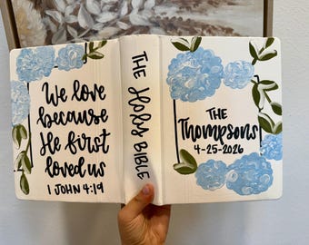 May include: A white Bible with a hand-painted cover featuring light blue hydrangea flowers and green leaves. The front cover displays the text "We love because He first loved us 1 JOHN 4:19" and the back cover says "THE THOMPSONS 4-25-2026".
