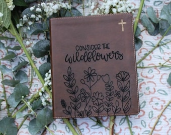 May include: A brown leather journal with the words "Consider the wildflowers" and a cross symbol. The journal features a floral design and is surrounded by greenery and white flowers.