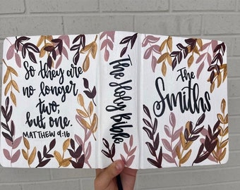 May include: A white Bible with a floral design in shades of brown, pink, and gold. The text "The Holy Bible" is written on the spine. The front cover has the text "The Smiths" and the verse "So they are no longer two, but one. Matthew 9:16".