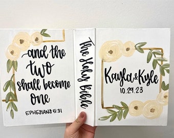 May include: A white canvas book with a gold frame and floral design. The text "The Holy Bible" is written on the spine. The front cover has the text "and the two shall become one Ephesians 5:31". The back cover has the text "Kayla & Kyle 10.29.23".