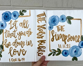 May include: A white wooden book with a gold inscription that reads "The Holy Bible" on the spine. The front cover features a gold frame with the text "Let all that you do be done in love 1 COR. 16:14" and blue floral designs. The back cover features a gold frame with the text "The Simpsons 11.11.23" and blue floral designs.