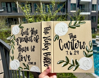 May include: A beige canvas book with white roses and green leaves painted on the cover. The text "The Holy Bible" is written on the left side of the book and "The Carters 7.6.24" is written on the right side. The left side of the book also has the text "...but the greatest of these is love 1 COR. 13" painted on it.