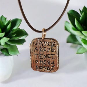 Può includere: Una collana con pendente color bronzo con l'iscrizione "SATOR AREPO TENET OPERA ROTAS". Il pendente rettangolare è appeso a un cordino marrone. Sullo sfondo, delle piante grasse verdi.