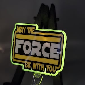 May include: A rectangular badge with the text "MAY THE FORCE BE WITH YOU" in white letters on a yellow and black background. The badge has a neon green border and a clear plastic clip.