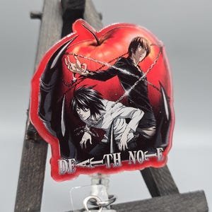 May include: A badge reel featuring anime characters within a red apple shape. The characters are depicted in a dynamic pose with chains. The text "DEATH NOTE" is printed across the bottom. The reel has a clear plastic holder and a silver clip.