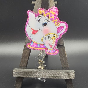 May include: A pink glitter-covered  retractable badge holder featuring a cartoon image of Mrs. Potts and Chip from Disney's Beauty and the Beast.
