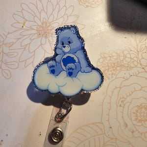 May include: A blue Care Bear with a blue heart on its chest sits on a white cloud with blue glitter. The Care Bear is wearing a blue headband and has a sad expression. The cloud is attached to a clear plastic retractable badge holder.