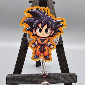 May include: A clear plastic badge reel with a round, orange and yellow glitter-filled charm featuring a cartoon character with black hair and an orange outfit. The character is wearing a blue belt and has a serious expression.