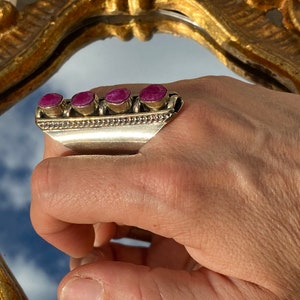 Può includere: Un anello d'argento con quattro pietre preziose rosa incastonate in fila. L'anello è progettato per essere indossato sul nodo delle dita.
