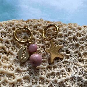 Könnte beinhalten: Ein Paar goldene Creolen mit Anhängern. Ein Ohrring hat eine rosa Steinperle und einen goldenen Anhänger mit dem Wort "Aloha" darauf. Der andere Ohrring hat einen goldenen Seestern-Anhänger.