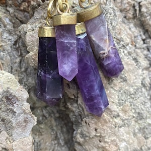 May include: Four amethyst crystal pendants with gold-toned metal settings. The crystals are faceted and have a purple hue. The pendants are hanging from a gold chain.