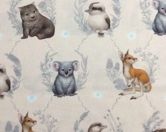 May include: A beige fabric with a repeating pattern of Australian animals, including a koala, a wombat, a kangaroo, a platypus, an echidna, and a kookaburra. The animals are surrounded by grey-green foliage and white flowers.