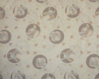 May include: A white fabric with a repeating pattern of brown circles containing a cartoon image of Dumbo the elephant. The background is a light beige with a scattered pattern of small brown leaves.