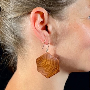 May include: A hexagonal dangle earring featuring a polished wood grain pattern with swirling light and dark brown tones. The earring is suspended from a thin silver-toned metal hook that passes through the earlobe, showcasing a natural wood texture.