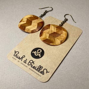 May include: A pair of dangle earrings featuring circular wooden pendants with a geometric parquet pattern in light and dark brown tones. The earrings hang from silver-toned metal hooks and are displayed on a tan card with the text Bush à Oreilles.