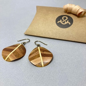 May include: A pair of drop earrings featuring a leaf-shaped design crafted from light and dark brown wood segments arranged in a herringbone pattern with a central cream-colored stripe and bronze-toned metal hooks, accompanied by a brown gift box.