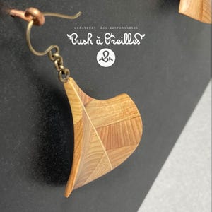 May include: A dangle earring featuring a curved, fan-shaped wooden pendant made of light and medium-toned wood segments. The earring hangs from a bronze-colored metal hook and chain. The text Bush à Oreilles is visible above.