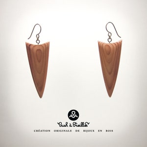 Puede incluir: Un par de pendientes de madera con forma de triángulo. Los pendientes están hechos de madera marrón claro y tienen una veta de madera natural. Los pendientes cuelgan de ganchos plateados.