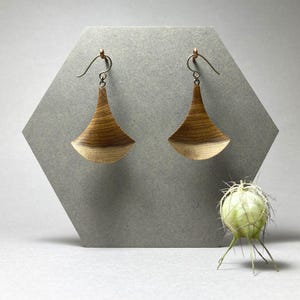 May include: A pair of dangle earrings featuring curved, fan-shaped wooden pendants with a natural wood grain pattern. The earrings are suspended from thin, metallic ear wires that attach to the top of each piece, showcasing a light to dark brown gradient.