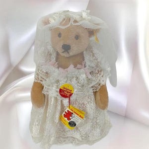 May include: A plush teddy bear dressed in a white lace wedding dress and veil. The bear has a tag that reads "Original" and "Für immer Dein". The dress features delicate lace detailing and a pink ribbon. The bear's fur is light brown.