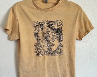May include: A light brown t-shirt with a black and white graphic print of a person's face with clouds around it.