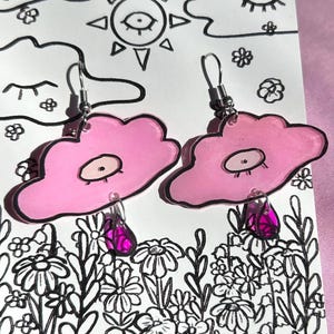 May include: Pink cloud shaped earrings with a single eye on each cloud and a pink teardrop hanging from each cloud.