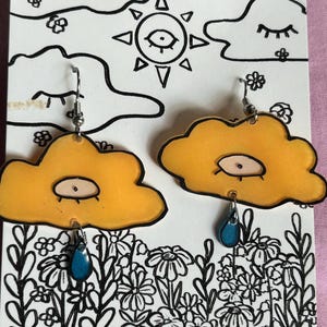 May include: A pair of yellow cloud-shaped earrings with a blue teardrop hanging from each. The clouds have a small eye drawn on them.