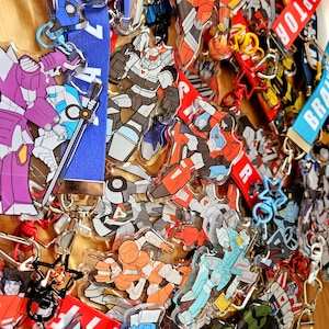 May include: A collection of acrylic keychains featuring various robot characters in vibrant colours like blue, purple, orange, red, and green. Each keychain is attached to a colourful fabric strap with text, including the word INTERCEPTOR.