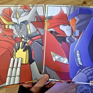 May include: Two Transformers robots, one red and one blue, are shown in a close-up image. The red robot is on the left and the blue robot is on the right. The red robot has a white and yellow face and the blue robot has a red face. Both robots are smiling.