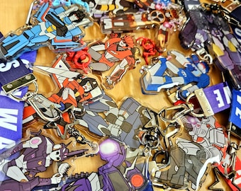 May include: A collection of acrylic keychains featuring various robot characters in vibrant colors, including red, blue, purple, and grey. Each keychain is attached to a metal clasp and rests on top of several fabric lanyards with printed text.