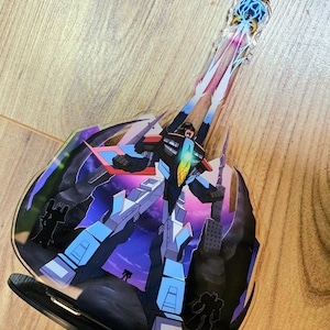 May include: A blue, red, and black robot character with a white and yellow energy blast coming from its hands. The robot is standing on a dark purple and blue background with a city skyline in the background. The robot is a character from the Transformers animated series.