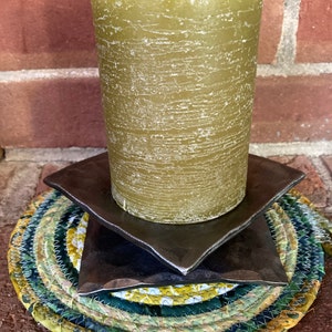 May include: A green pillar candle sits on a stack of three dark brown square coasters. The coasters are on a round, multi-colored braided rug.