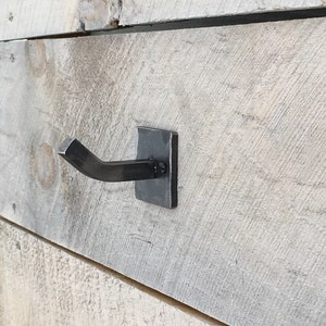 May include: A black metal hook mounted on a wooden wall. The hook is bent at a 90-degree angle and has a square base.