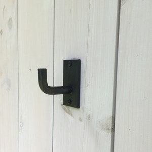 May include: Black metal hook mounted on a white wooden wall. The hook is shaped like a 'J' and has a flat back plate.