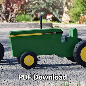 May include: A green wooden tractor with black tyres and yellow centres. The tractor features a yellow stripe and the words "JOHN DEERE" and "2955". A black vertical pole is on top of the tractor.