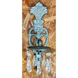 May include: A silver-toned metal wall sconce featuring an ornate, symmetrical backplate with intricate scrollwork and floral motifs. A circular candle holder platform is attached to the base, adorned with multiple clear, faceted glass teardrop crystals.