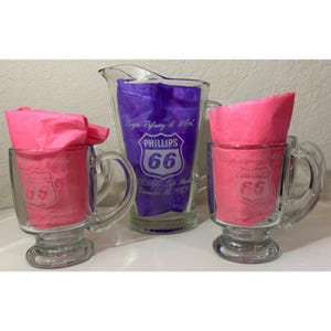 May include: A clear glass pitcher and two matching glass mugs, each featuring the Phillips 66 logo. The pitcher is etched with Borger Refinery & NGL, 100,000 Safe Manhours, December 16, 1983. Bright pink tissue paper is tucked inside each glass vessel.