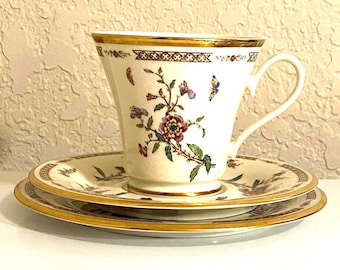May include: A cream-colored porcelain teacup and saucer set featuring a delicate floral pattern with small butterflies, accented by a gold rim and a geometric border design along the top edge of the cup and the outer rim of the saucer.
