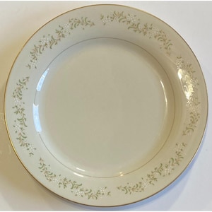 May include: Cream-colored ceramic plate with a gold rim and delicate floral pattern. The floral design features small white flowers and green leaves. A thin gold line separates the floral border from the center.