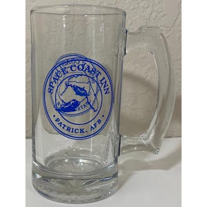 May include: A clear glass beer mug featuring a blue circular graphic with the text SPACE COAST INN and PATRICK, AFB. The design includes a map outline and a space shuttle illustration. The mug has a thick handle and a starburst pattern on the base.