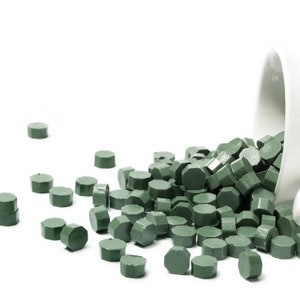 May include: A white ceramic container spilling out green wax sealing pellets. The pellets are hexagonal in shape and are scattered on a white surface.