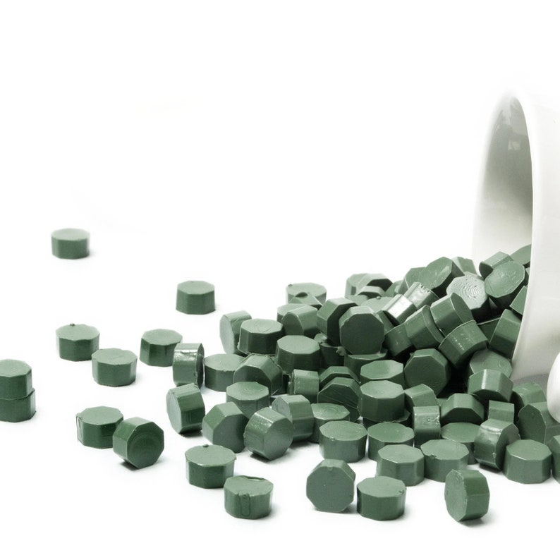 May include: A white ceramic container spilling out green wax sealing pellets. The pellets are hexagonal in shape and are scattered on a white surface.