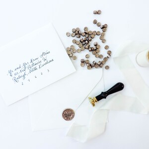 May include: A white envelope with a handwritten address, a spool of white ribbon, a wax seal stamp, and a pile of wax seal pellets. The address reads "Mr. and Mrs. Krew Stone, 123 New Biltmore W, Raleigh, North Carolina 27603".