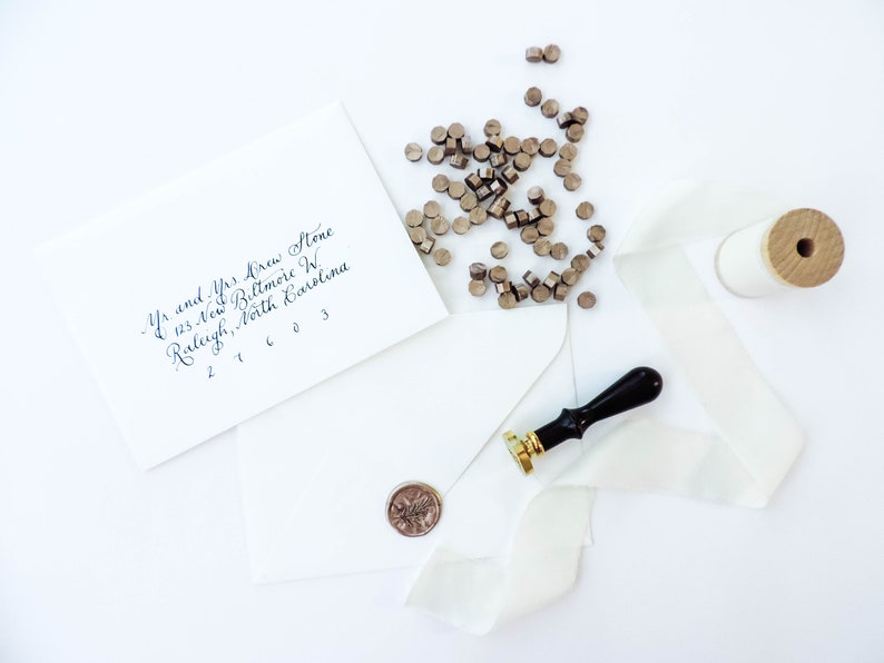 May include: A white envelope with a handwritten address, a spool of white ribbon, a wax seal stamp, and a pile of wax seal pellets. The address reads "Mr. and Mrs. Krew Stone, 123 New Biltmore W, Raleigh, North Carolina 27603".