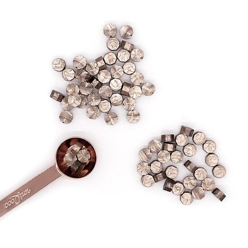 May include: A close-up of a pile of silver-colored wax seals, with a few more in a small copper measuring spoon. The spoon has the word "cookspoon" printed on the handle.