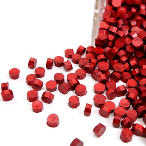 May include: Red sealing wax beads in a clear container, spilling out onto a white surface. The beads are small and round, with a metallic sheen.