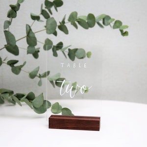 May include: Clear acrylic table number sign with the text "TABLE two" in white calligraphy on a wooden stand.