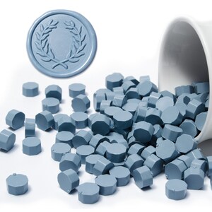 May include: A close-up of a blue wax seal with a laurel wreath design. A white ceramic container is spilling out a large quantity of blue wax seal pellets.