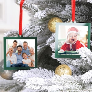 May include: Two square glass Christmas ornaments with green frames. One ornament features a family photo of seven people, and the other ornament features a photo of a baby wearing a red Santa hat.